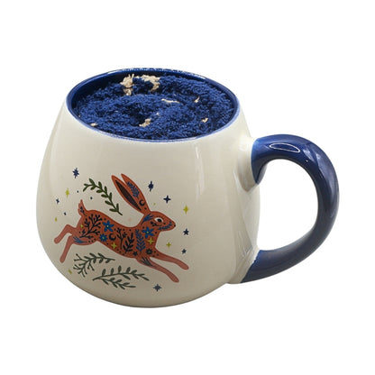 A ceramic mug with a cream-colored exterior, a blue handle and interior, and a painted brown rabbit design decorated with blue and green details on the front.