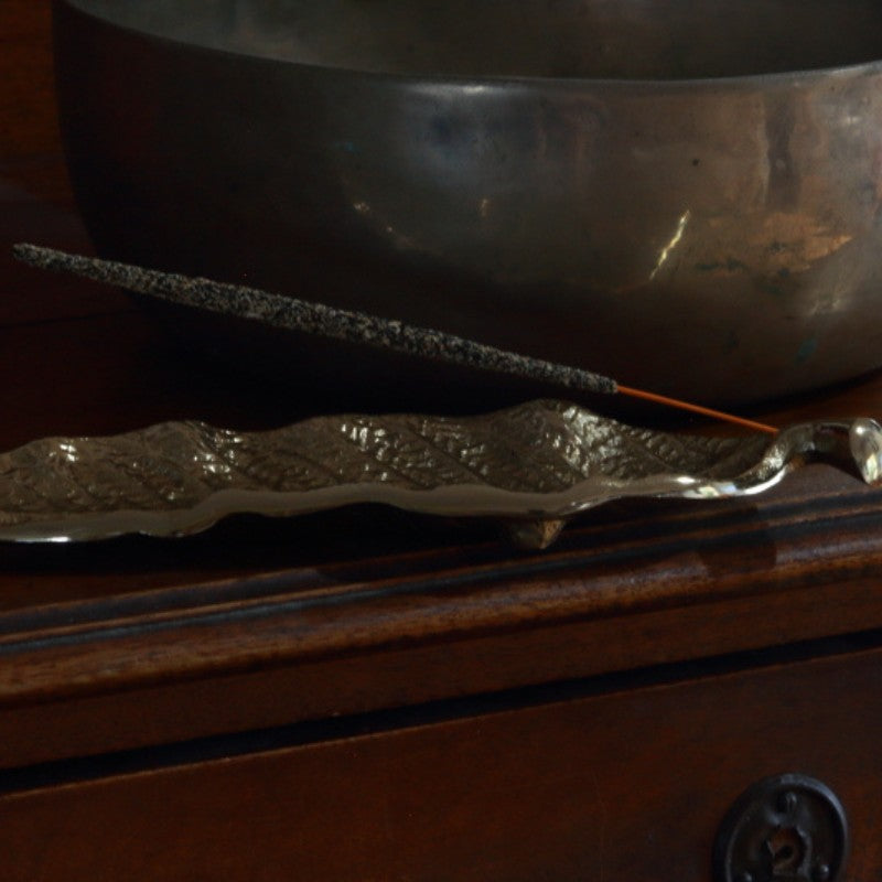 gold incense holder in the shape of a leaf holding a natural incense stick , on a wooden sideboard, in front of a singing bowl