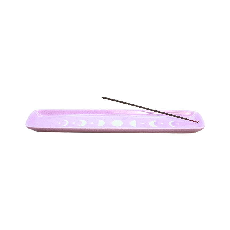 A lavender purple rectangular ceramic incense holder with white moon phase cutouts, featuring a single dark incense stick resting diagonally across the top, set against a solid white background.
