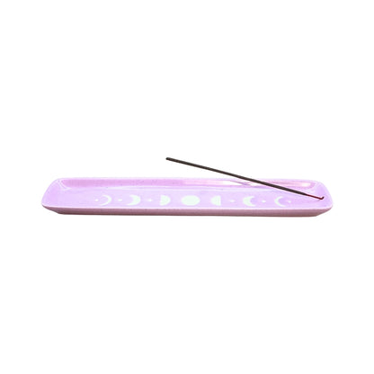 A lavender purple rectangular ceramic incense holder with white moon phase cutouts, featuring a single dark incense stick resting diagonally across the top, set against a solid white background.