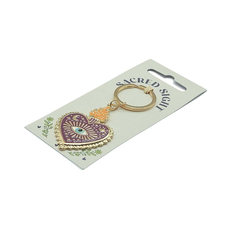 heart shaped gold and purple key ring with an eye motif, on a cardboard backing, in front of a white background