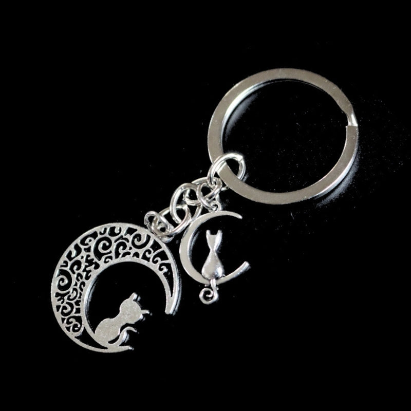silver coloured key ring with a a large and small crescent moon each with a cat sitting on the moon,   joined to a silver chain which links onto a silver ring on a black background