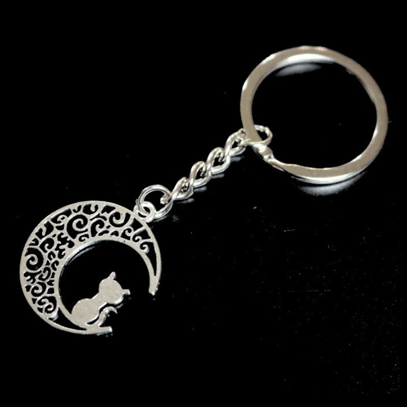 silver coloured key ring with a cat sitting on a rescent moon  joined to a silver ring by a silver chain on a black background