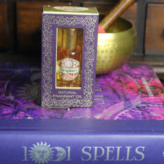 purple and gold box containing a miniature perfume bottle, sitting on a purple and pink book of spells in front of a brass singing bowl with wooden striker. in the background there is an apothecary cabinet