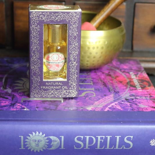 purple and gold box containing a miniature perfume bottle, sitting on a purple and pink book of spells in front of a brass singing bowl with wooden striker. in the background there is an apothecary cabinet