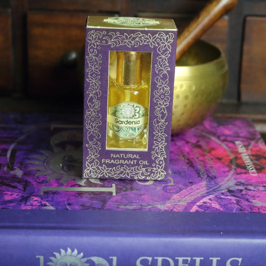 purple and gold box containing a miniature perfume bottle, sitting on a purple and pink book of spells in front of a brass singing bowl with wooden striker. in the background there is an apothecary cabinet