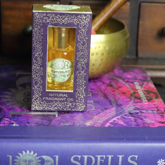 purple and gold box containing a miniature perfume bottle, sitting on a purple and pink book of spells in front of a brass singing bowl with wooden striker. in the background there is an apothecary cabinet