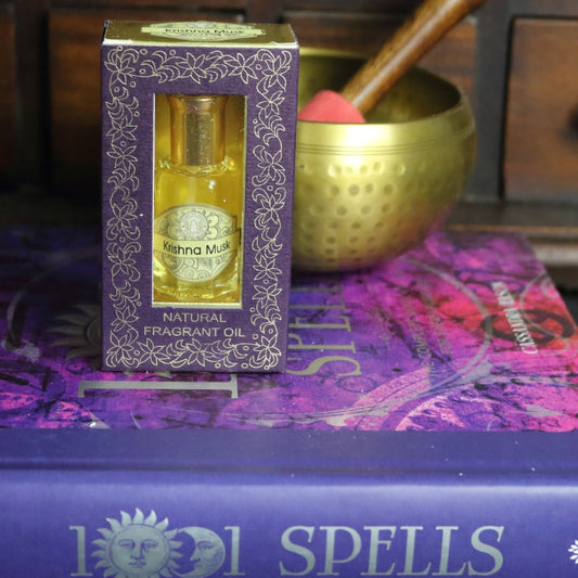 purple and gold box containing a miniature perfume bottle, sitting on a purple and pink book of spells in front of a brass singing bowl with wooden striker. in the background there is an apothecary cabinet