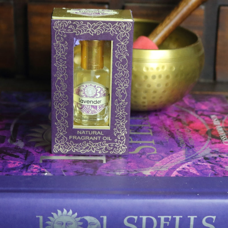 purple and gold box containing a miniature perfume bottle, sitting on a purple and pink book of spells in front of a brass singing bowl with wooden striker. in the background there is an apothecary cabinet