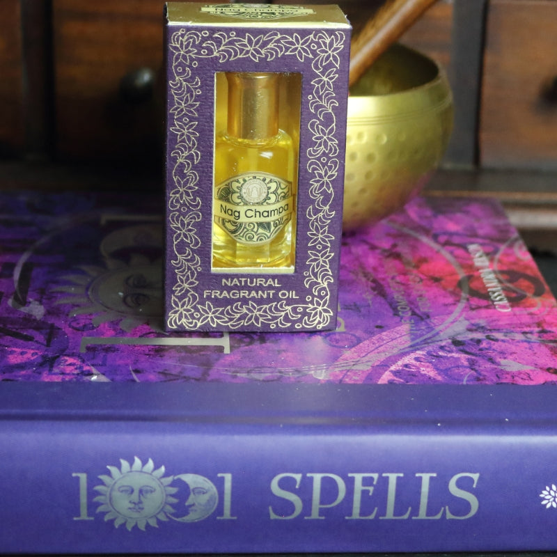 purple and gold box containing a miniature perfume bottle, sitting on a purple and pink book of spells in front of a brass singing bowl with wooden striker. in the background there is an apothecary cabinet