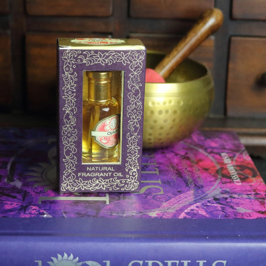 purple and gold box containing a miniature perfume bottle, sitting on a purple and pink book of spells in front of a brass singing bowl with wooden striker. in the background there is an apothecary cabinet