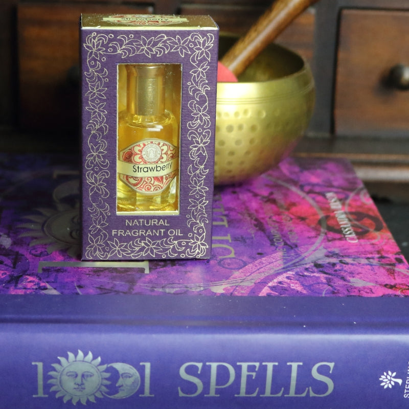 purple and gold box containing a miniature perfume bottle, sitting on a purple and pink book of spells in front of a brass singing bowl with wooden striker. in the background there is an apothecary cabinet