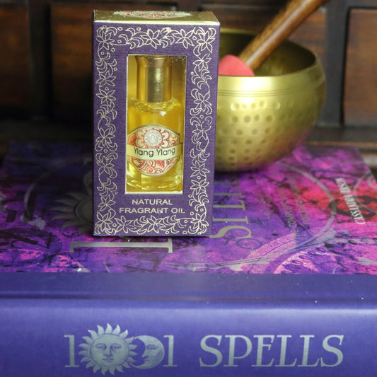 purple and gold box containing a miniature perfume bottle, sitting on a purple and pink book of spells in front of a brass singing bowl with wooden striker. in the background there is an apothecary cabinet