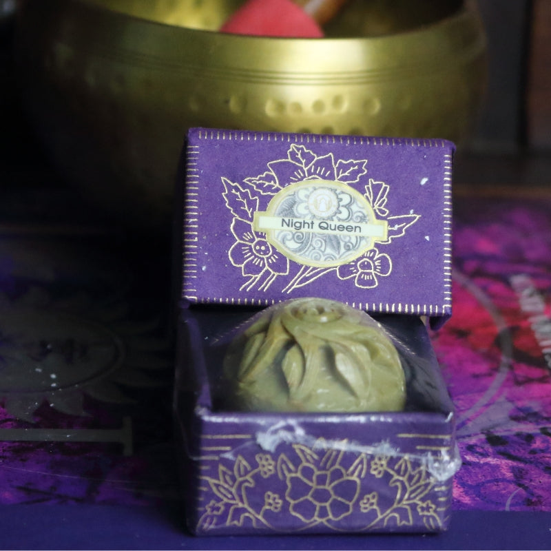 purple and gold box containing a hand carved soapstone jar, sitting on a purple and pink book of spells in front of a brass singing bowl with wooden striker.