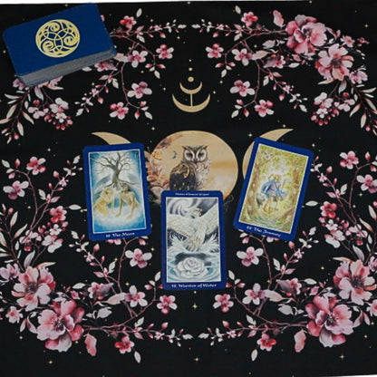 black tarot cloth with gold triple moon and owl design with floral borders.  3 card spread is laid out on top and a tarot deck in the corner