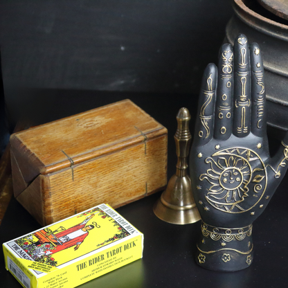 rider waite tarot deck sitting in front of a wooden tarot box next to an old cauldron, brass altar bell and black carved hand with gold symbols on its palm.