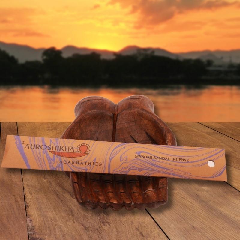 pk of auroshikha brand incense sticks sitting on a carved wooden open hand, on a wooden jetty in front of a sunset sky