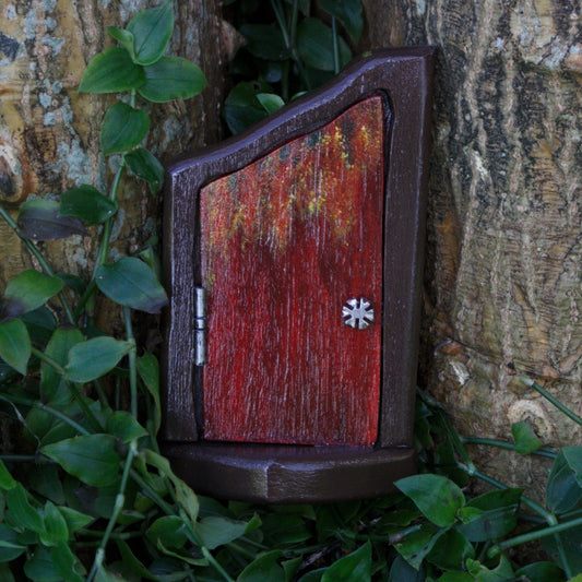 red and brown autumn fairy door with silver hinge and snowflake silver door knob in front of a tree and greenery