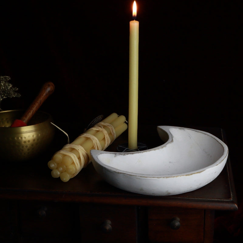 lit beeswax prayer candle behind a whitewashed crescent moon carved wooden bowl and brass singing bowl, sitting an a wooden apothecary cabinet