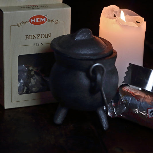 black cast iron cauldron in front of a beige coloured box of benzoin resin, next to a lit white church candle and a silver roll of charcoal discs and pair of stainless steel charcoal tongs on a wooden table