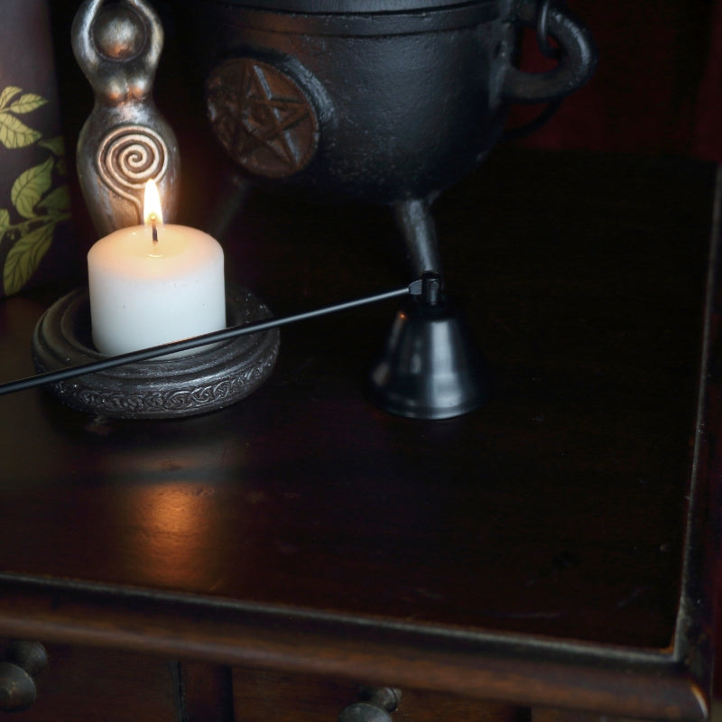 apothecary cabinet with a spellcrafting book on top, a black cast iron cauldron with a pentcle design on front, a black candle snuffer, candle holder in the shape of a goddess holding a triple moon.