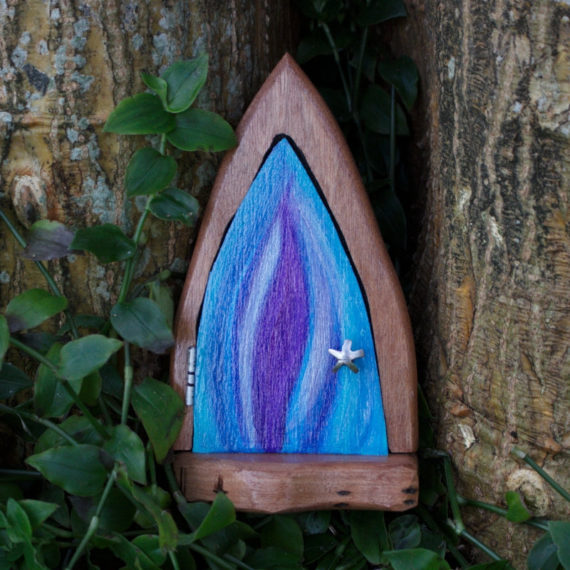 wooden fairy door with blue silver and purple flame door, silver starfish doorknob and silver hinge, leaning against a tree with greenery underneath