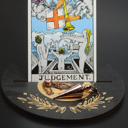 bronze conical pendulum sitting on a moon shaped piece of wood painted black with a crescent moon on it, with a groove cut down the middle of it, holding a judgement rider waite tarot card.
