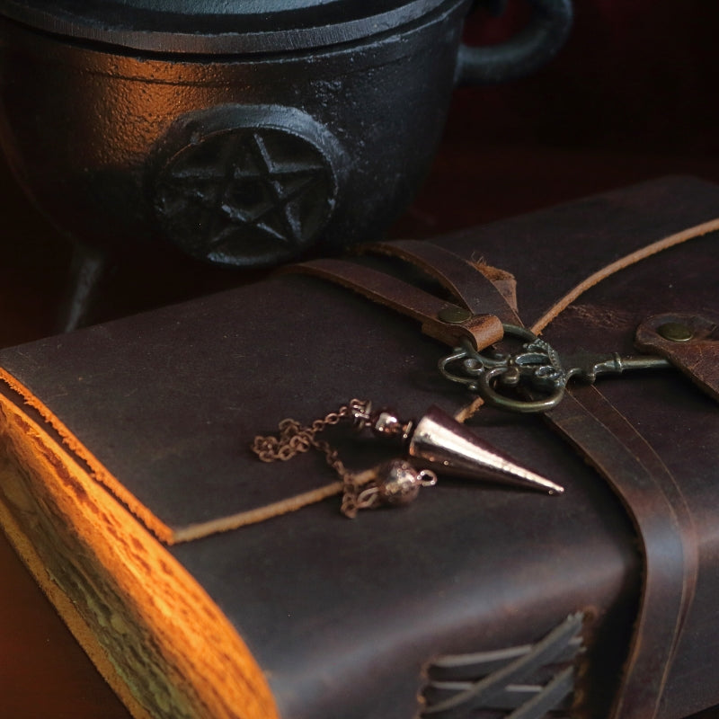 bronze conical pendulum sitting on a brown leather journal, in front of a black cast iron cauldron with a pentacle symbol on the front