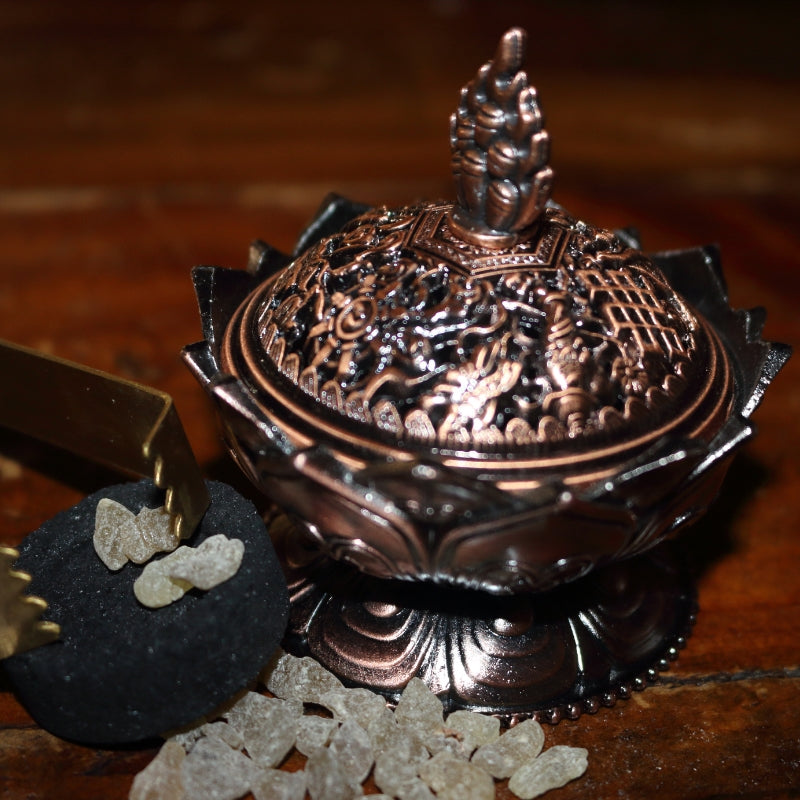 ornate red brass coloured 2 piece censer/ loose incense burner on a wooden table, next to a charcoal disc and pair of gold charcoal tongs with frankincense granules strewn around the base of it.