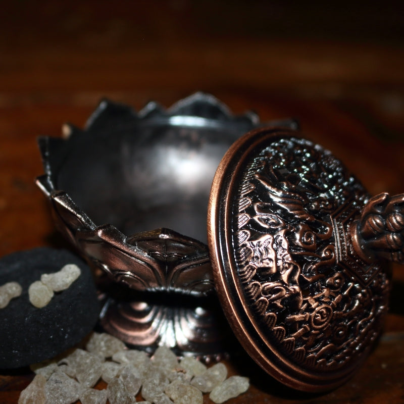 ornate red brass coloured 2 piece censer/ loose incense burner sitting open on a wooden table, next to a charcoal disc and pair of gold charcoal tongs with frankincense granules strewn around the base of it.