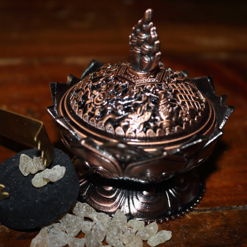 ornate red brass coloured 2 piece censer/ loose incense burner on a wooden table, next to a charcoal disc and pair of gold charcoal tongs with frankincense granules strewn around the base of it.