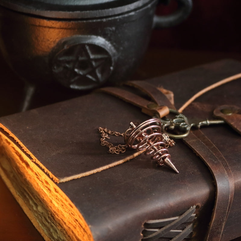 bronze spiral pendulum sitting on a brown leather journal, in front of a black cast iron cauldron with a pentacle symbol on the front