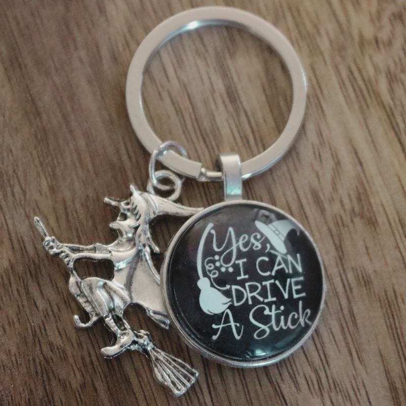 keyring of a witch riding a broomstick, with a black circle attached to it saying "yes i can drive a stick" in white writing on a black background