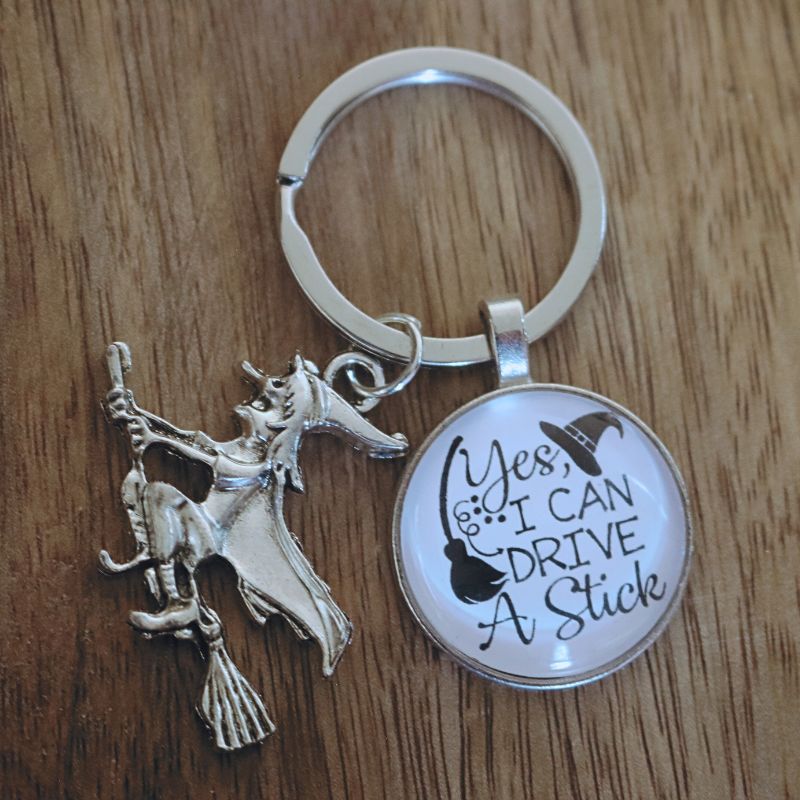 keyring of a witch riding a broomstick, with a black circle attached to it saying "yes i can drive a stick" in black writing on a white background