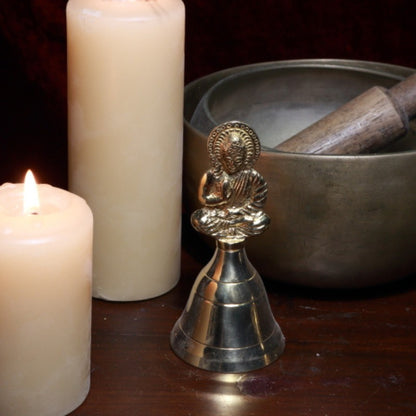 brass bell with Buddha on handle next to beeswax candles and singing bowls