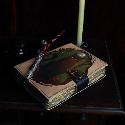 leather bound book with an image of a black cat on the front. A red feather pen sitting on top of the book, in front of a beeswax candle