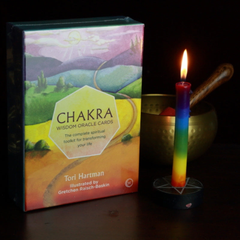 rainbow spell candle in a pentacle candle holder in front of a brass singing bowl, next to a pack of chakra oracle cards