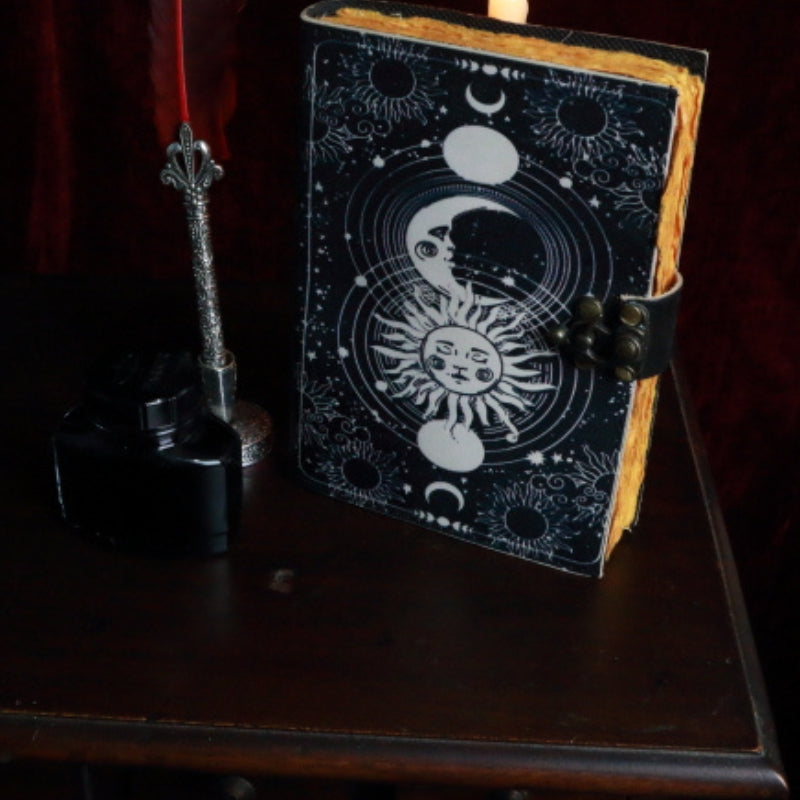 leather bound book with an image of a sun and moon on the front. A red feather pen and ink  sitting next to the book,