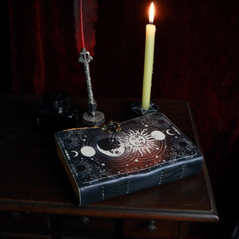 leather bound book with an image of a sun and moon on the front. A red feather pen sitting next to the book, in front of a beeswax candle