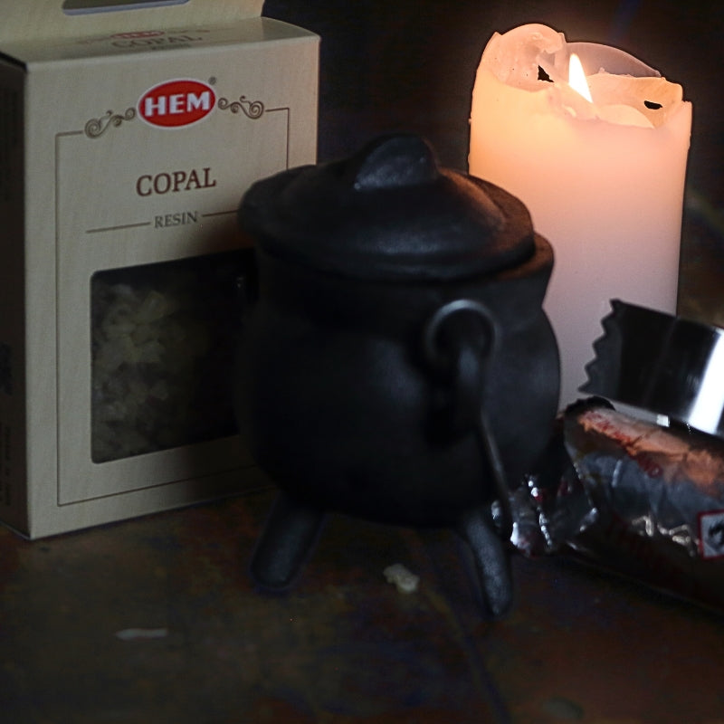 black cast iron cauldron in front of a beige coloured box of copal resin, next to a lit white church candle and a silver roll of charcoal discs and pair of stainless steel charcoal tongs on a wooden table