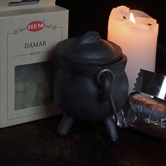 black cast iron cauldron in front of a beige coloured box of damar blood resin, next to a lit white church candle and a silver roll of charcoal discs and pair of stainless steel charcoal tongs on a wooden table