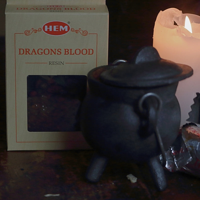 black cast iron cauldron in front of a beige coloured box of dragon's blood resin, next to a lit white church candle and a silver roll of charcoal discs and pair of stainless steel charcoal tongs on a wooden table