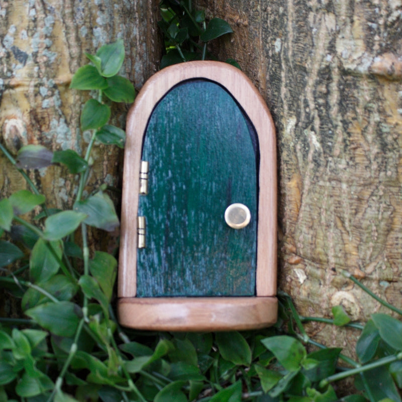 handmade wooden fairy door with a green door and handmade brass hinges and doorknob, sitting between trees and on top of dark green vines