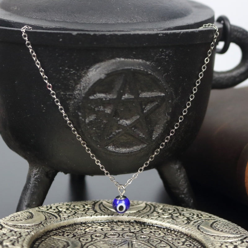 silver necklace with a blue, white and black eye shaped bead. The necklace is wrapped around a cast iron cauldron with pentacle symbol on the front, above an ornate silver plate with moons embossed on it.