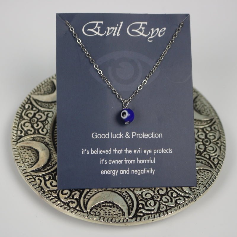 silver necklace with a blue, white and black eye shaped bead, tied to a blue cardboard backing with the evil eye symbol  on it. The necklace is sitting on an ornate silver plate with moons embossed on it.