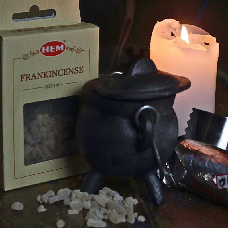 black cast iron cauldron in front of a beige coloured box of frankincense resin, with frankincense granules in the foreground next to a lit white church candle and a silver roll of charcoal discs and pair of stainless steel charcoal tongs on a wooden table
