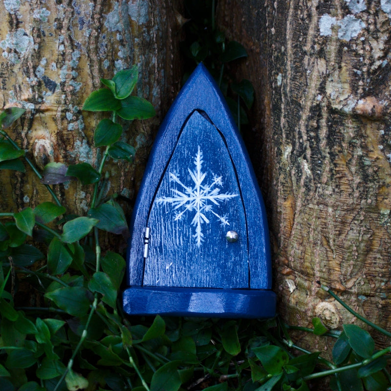 Blue Fairy Door with a white snowflake painted on the front in front of a tree