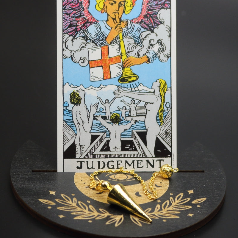 gold conical pendulum sitting on a moon shaped piece of wood painted black with a crescent moon on it, with a groove cut down the middle of it, holding a judgement rider waite tarot card.