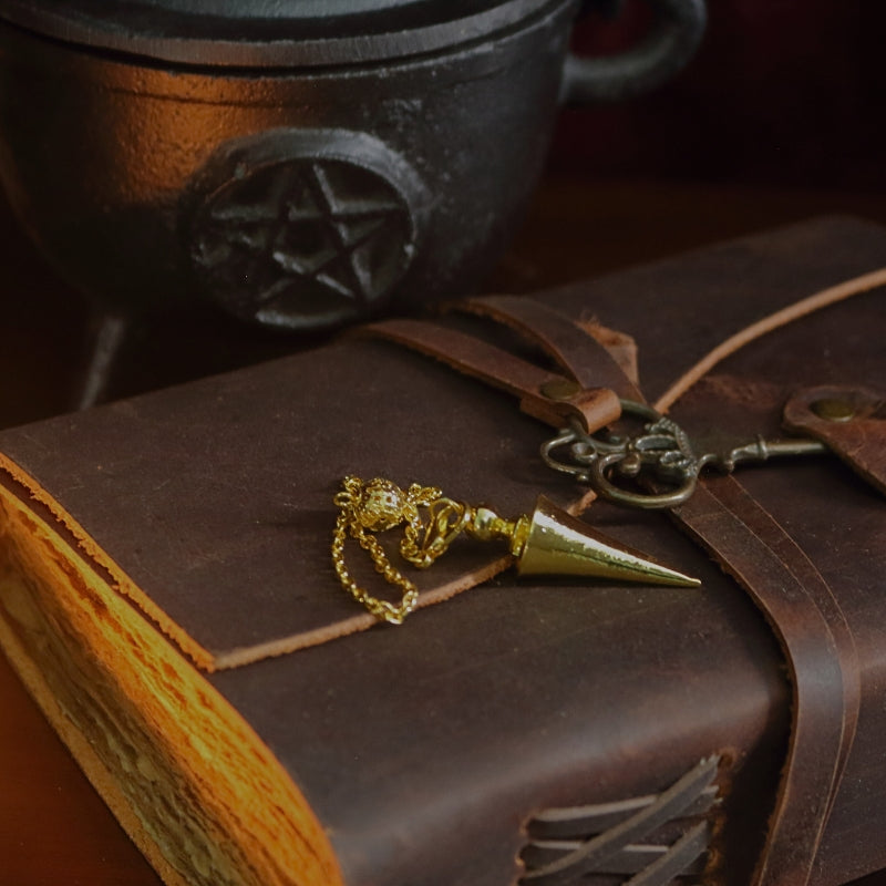 gold conical pendulum sitting on a brown leather journal, in front of a black cast iron cauldron with a pentacle symbol on the front 