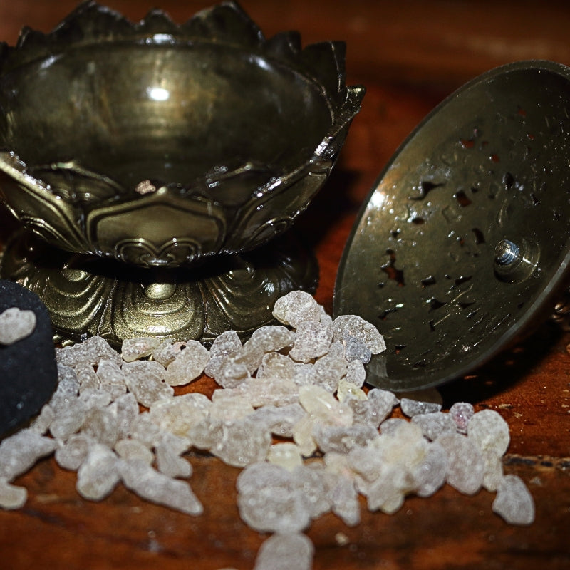 2 piece (top and bottom) ornate gold coloured incense holder, sitting  open on a wooden table, next to a charcoal disc, with frankincense granules strewn around the base of the incense holder